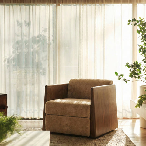 A boxy, low-profile armchair with thick wood side panels and textured brown upholstery set against a sunlit window.