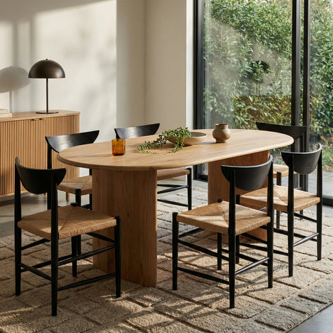 A minimalist dining room featuring a light wood oval table surrounded by six black chairs with woven seats on a textured rug.
