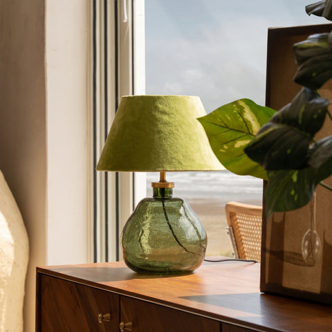 Green glass lamp on a wooden table near a bright window and houseplants.