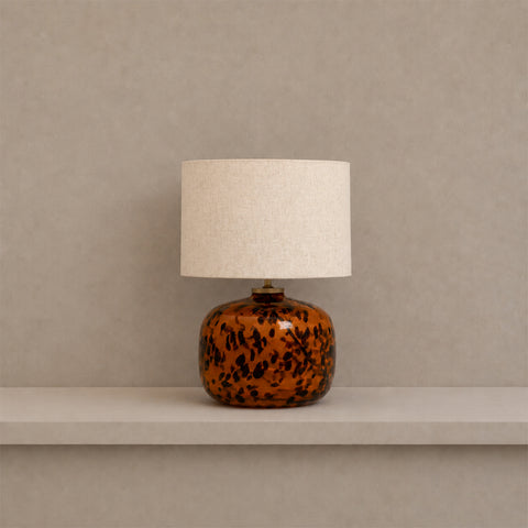 Tortoiseshell glass table lamp with a white drum shade sitting on a shelf.