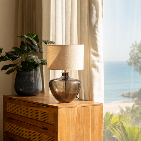Smoky glass lamp styled on a wooden dresser near a bright window with a coastal view.