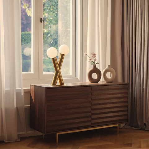 A modern gold table lamp with two frosted globe bulbs styled on a dark wood fluted sideboard next to decorative vases and a window.