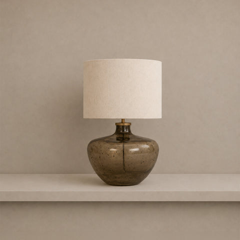 Smoky glass table lamp with a white drum shade sitting on a shelf.