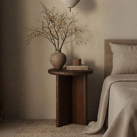 Wooden side table beside a bed, styled with a ceramic vase and dried branches in a calm, neutral bedroom.