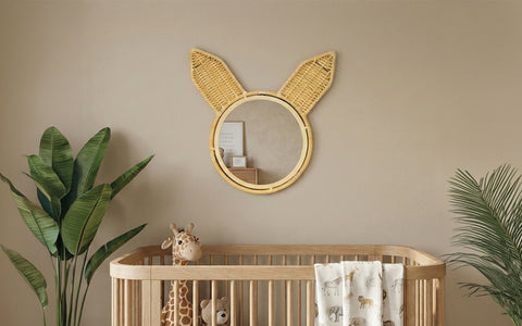 A whimsical round wall mirror with woven rattan bunny ears mounted above a wooden baby crib.