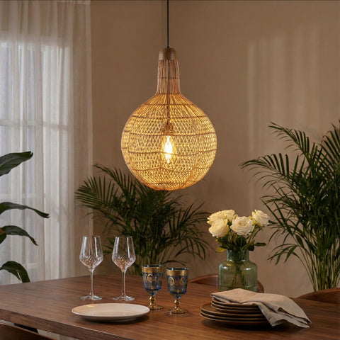 A large, round woven rattan pendant light illuminated over a dining table set with wine glasses and flowers.