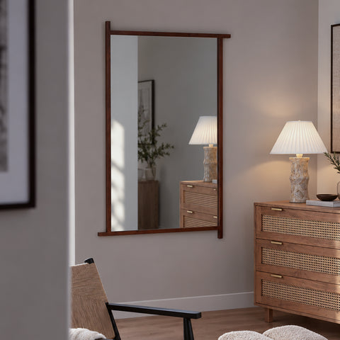 A tall rectangular wall mirror with a dark brown wooden frame hangs on a beige wall in a styled room. The mirror reflects a matching wooden chest of drawers and a lamp, while the room also features a wooden dresser with woven cane panels, a lit table lamp, and a modern chair.