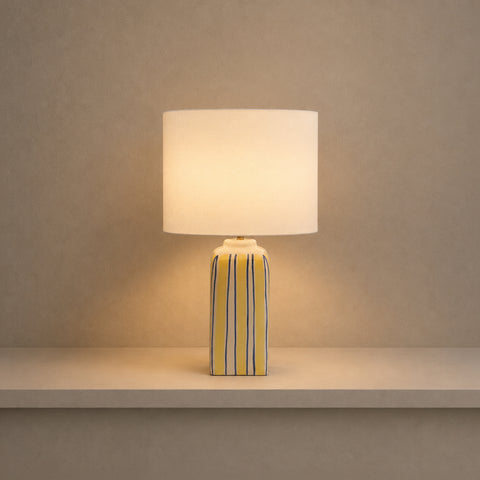 Illuminated striped table lamp glowing warmly on a shelf in a dim room.