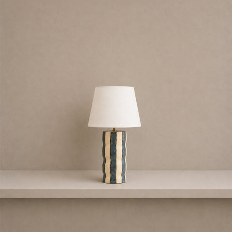 Small cylindrical patterned table lamp with a white tapered shade on a shelf.